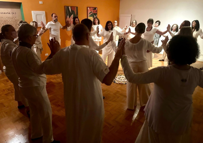 Roda meditativa: participantes vestidos de branco, com os braços erguidos, numa sala de parede laranja