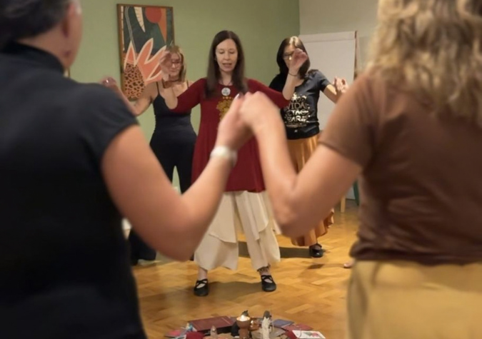 Mulheres de mãos dadas, com os braços erguidos, em roda em torno de uma pequena mandala de objetos no chão