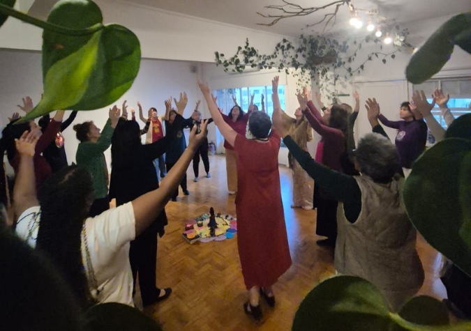 Roda de mulheres com os braços erguidos numa sala, vista entre as folhas de uma planta