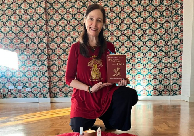 Estela Gomes ajoelhada, sorrindo, segurando o livro Mulheres que Correm com os Lobos, com uma vela diante dela