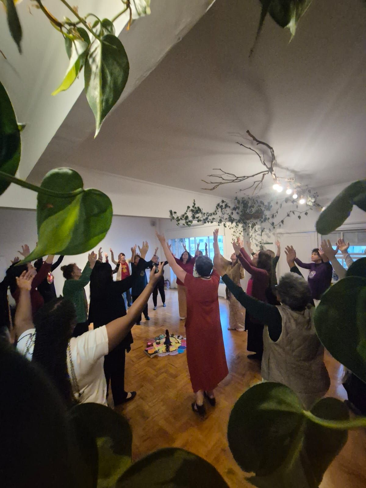 Roda de mulheres com os braços erguidos, vista entre as folhas de uma planta