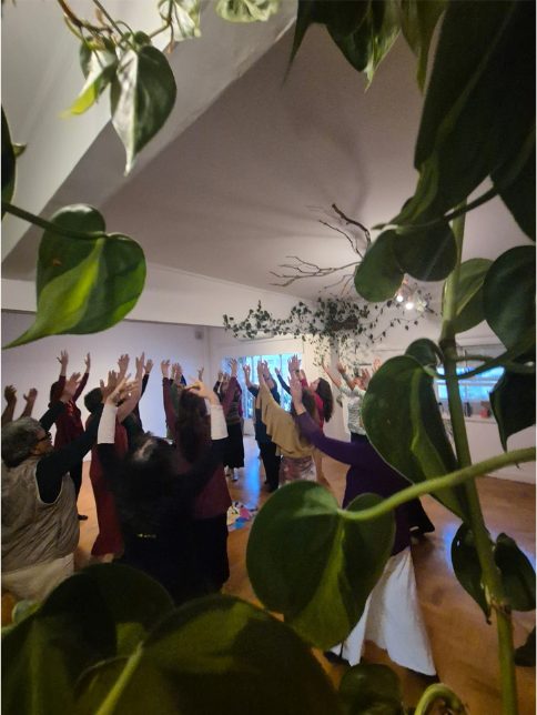 Mulheres com os braços erguidos dançando, vistas entre as folhas de uma planta